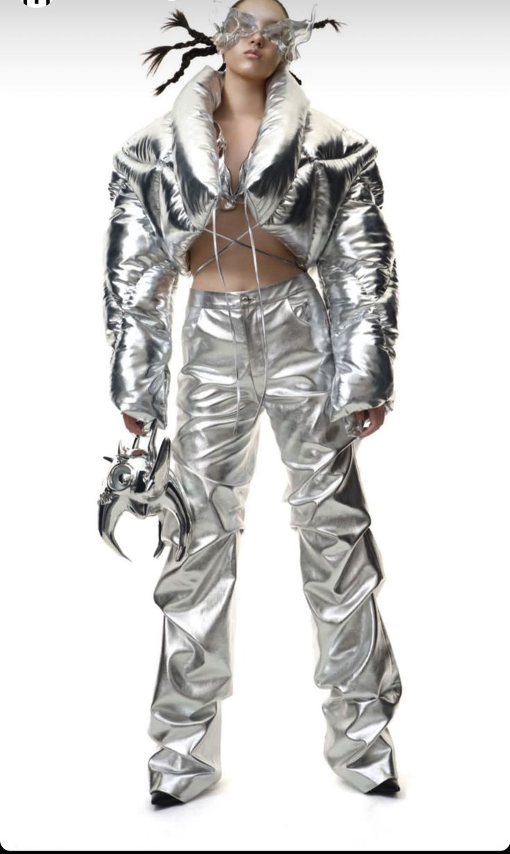 a futuristiclooking woman in a silver, metallic outfit. the outfit consists of a puffer jacket and pants. the jacket is cropped and has a ruffled collar. the pants are loosefitting and cinched at the waist. the woman is wearing silver, platform boots. she is standing with her arms by her side and is looking at the camera.