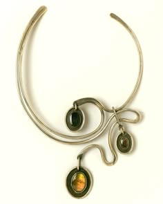 a silver necklace with three pendants. the necklace is a thick, open circle. the pendants are attached to the circle with thin silver chains. the pendants are oval shaped with gemstones set inside. the first pendant has a black gemstone, the second has a green gemstone, and the third has an orange gemstone. the necklace is a modern and elegant design.
