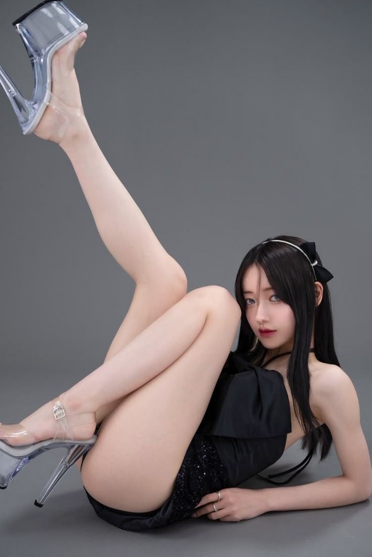 a woman is lying on the floor with one leg raised in the air. she is wearing clear high heels and a black dress. she has long black hair and is looking at the camera. the background is gray.