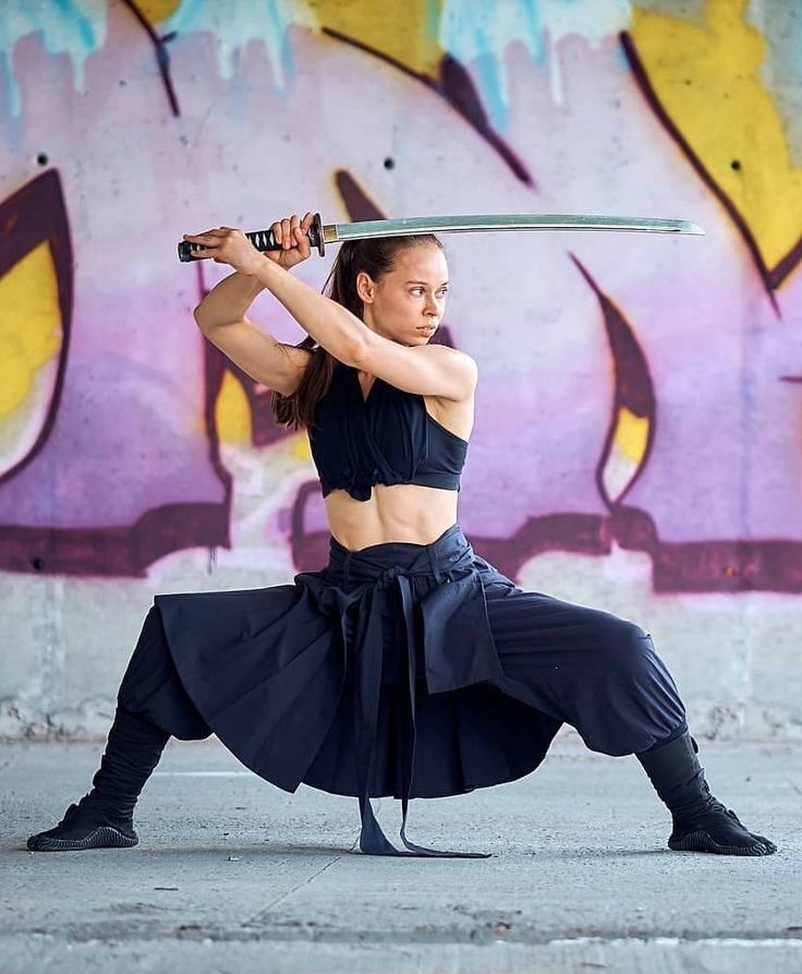 a woman dressed as a ninja holds a katana sword in a fighting stance. she is posed in front of a graffiti covered wall.