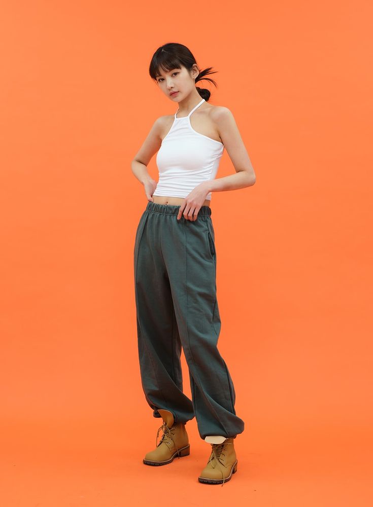 the image shows a young asian woman posing against a vibrant orange background. she's wearing a white ribbed halter top paired with loose, cuffed green pants and brown ankle boots. her hair is pulled back in a simple updo. the overall style is casual and comfortable yet fashionable. the bright background contrasts nicely with her outfit, making the clothing details and the model's pose stand out.