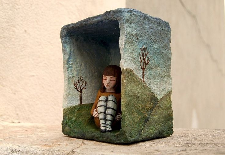 a handcrafted sculpture featuring a girl with brown hair and striped tights sitting inside a stonelike structure. the structure is painted to resemble a landscape with trees and a sky. the girl has her eyes closed, giving off a melancholic and introspective mood. the artwork evokes a feeling of loneliness and being sheltered in a personal world.