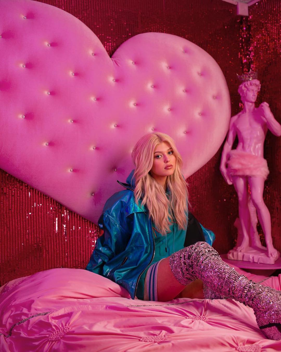 a young woman with blonde hair is sitting on a pink bed with a heartshaped headboard. she's wearing a blue jacket and sparkly silver thighhigh boots. behind her is a pink sequin wall and to her right is a pink statue of a man wearing a tiara and a furry skirt. the scene is very pink and bright.