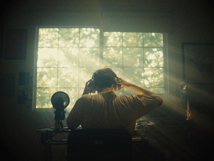 the image shows a person sitting in front of a microphone in a dimly lit room, bathed in sunbeams streaming through a large window.  the individual is wearing headphones and appears to be recording music or engaged in a similar audio production process. the scene is set in a home studio with a vintage or retro feel, emphasized by the film grain and muted colors. the back of the person is visible, emphasizing their focus on the task at hand. the overall mood is serene and reflective.