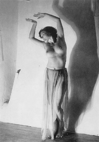 a black and white photograph depicts a nude female dancer posing against a white wall.  her arms are raised and her body is slightly arched, creating a graceful and elegant pose.  a significant portion of her body is obscured by a long, sheer skirt. her shadow is cast prominently on the wall behind her, adding depth and contrast to the image. the overall mood is artistic and timeless.