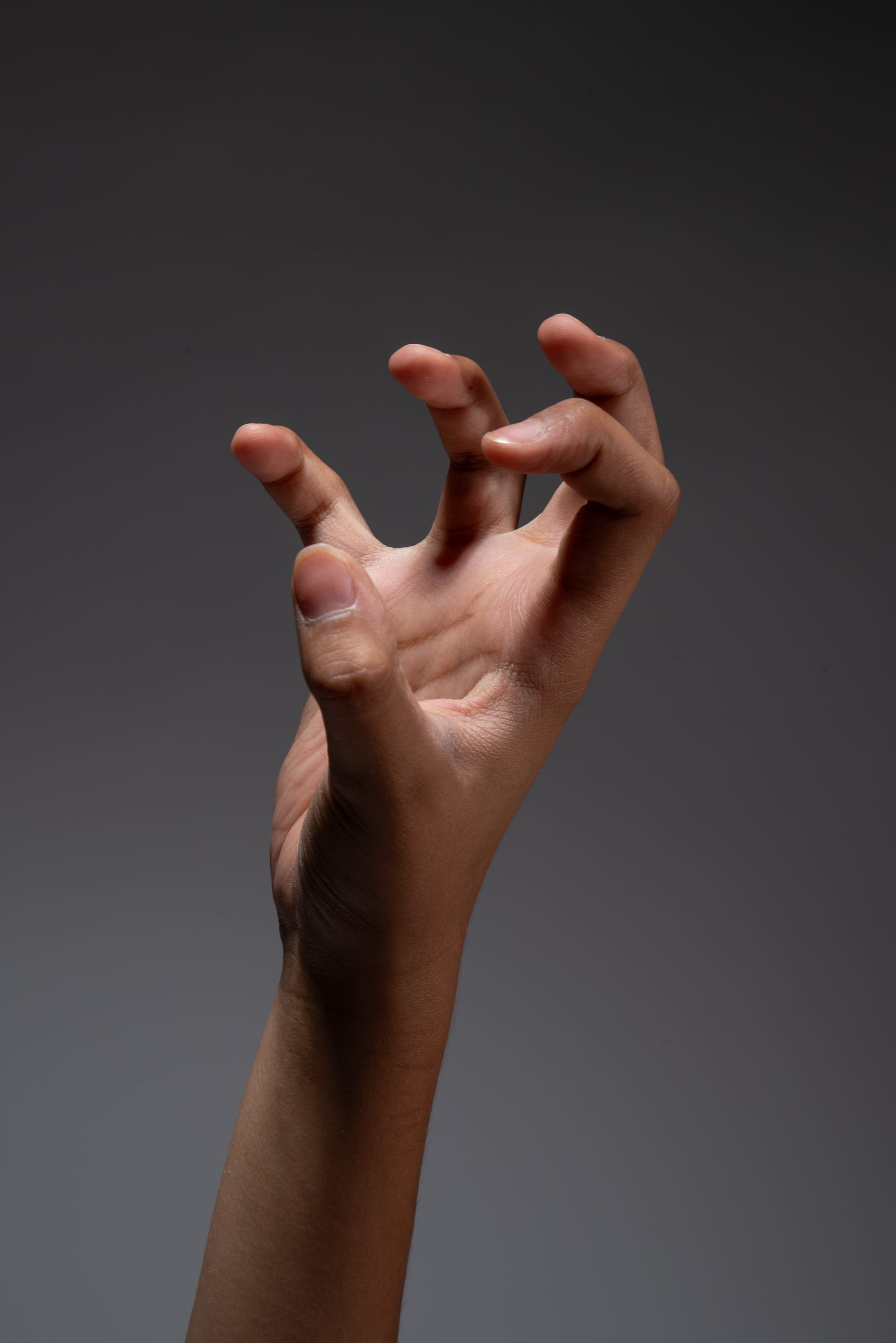 a hand with light brown skin tone is reaching up with its fingers curved in a clawlike gesture against a gray background.