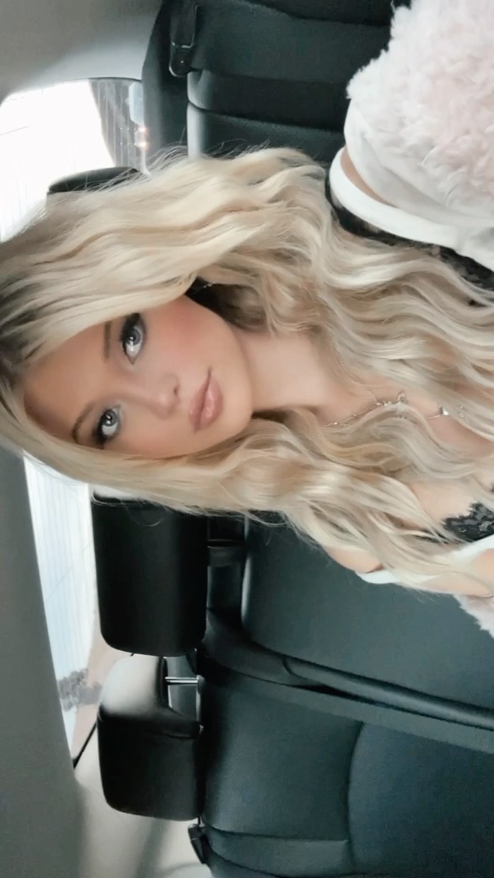 a selfie of a blonde woman in a car. she is wearing makeup and a necklace. she is sitting in a black leather car seat and wearing a white faux fur jacket.