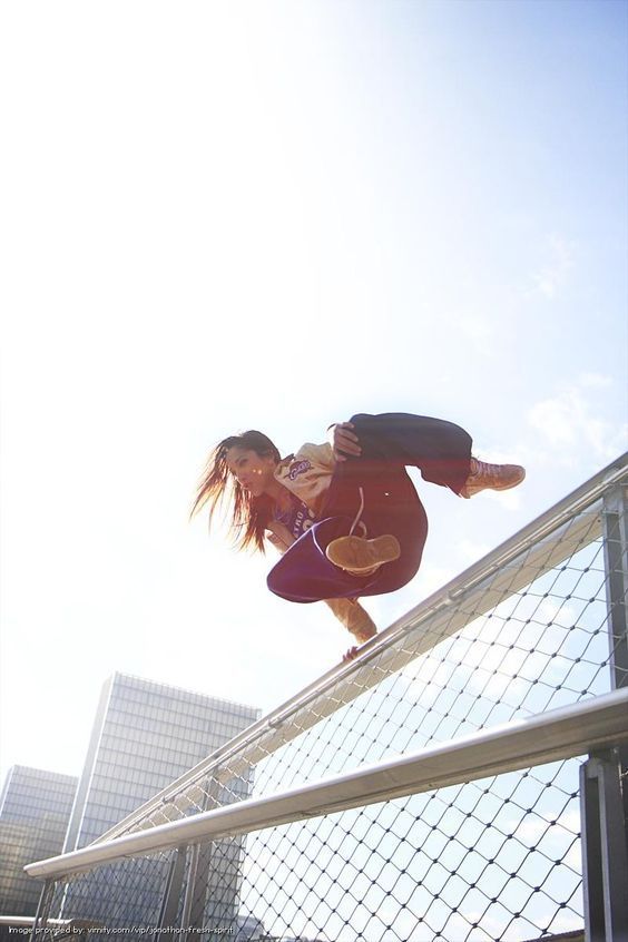 a young woman is seen jumping over a fence. she is wearing a jacket, black pants and sneakers. her hair is long and brown, she is wearing a pink wristband and a ring. the  fence is  made of wire mesh and there are buildings behind the fence. the sky is light blue.  this image shows a woman engaged in parkour or freerunning.  she is wearing sporty and comfortable clothes. she looks very athletic.  this is a photo of an adventurous and active woman outdoors.