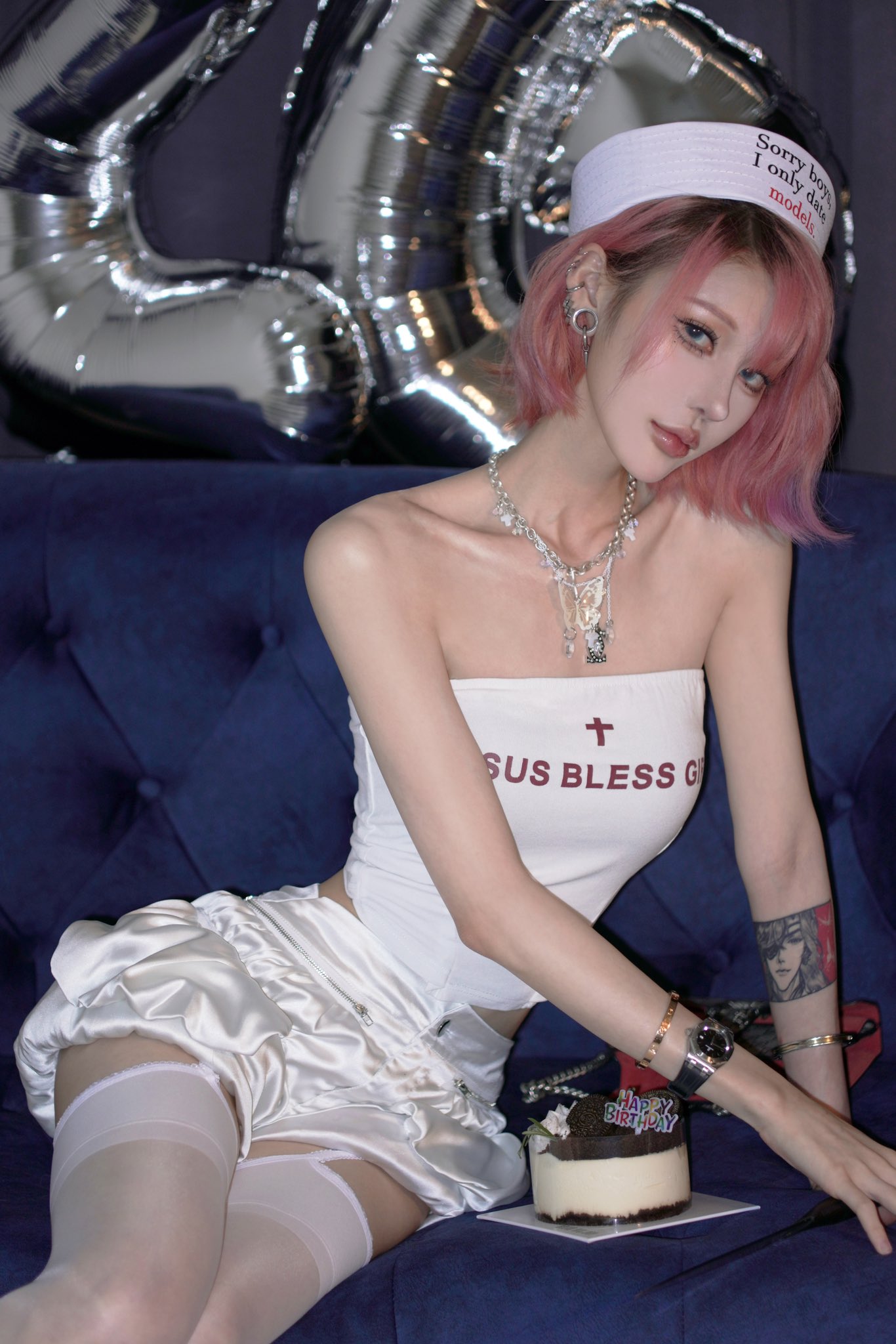 a young woman with pink hair is sitting on a blue couch. she is wearing a white nurse's hat with the words "sorry boys i only date models" on it, a white satin crop top that says "jesus bless g" and a white satin mini skirt. she is also wearing a necklace, bracelets and a watch.  there is a birthday cake in front of her and silver balloons are blurred in the background. she is posing for the camera and looks sexy and attractive.