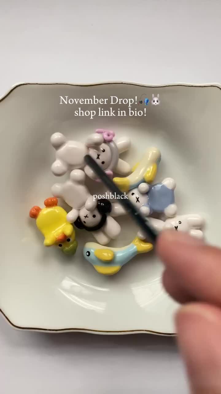 a bowl filled with various jewelry charms, including a duck, lambs, fish, and bunny. a hand is holding a black stick in the bowl.