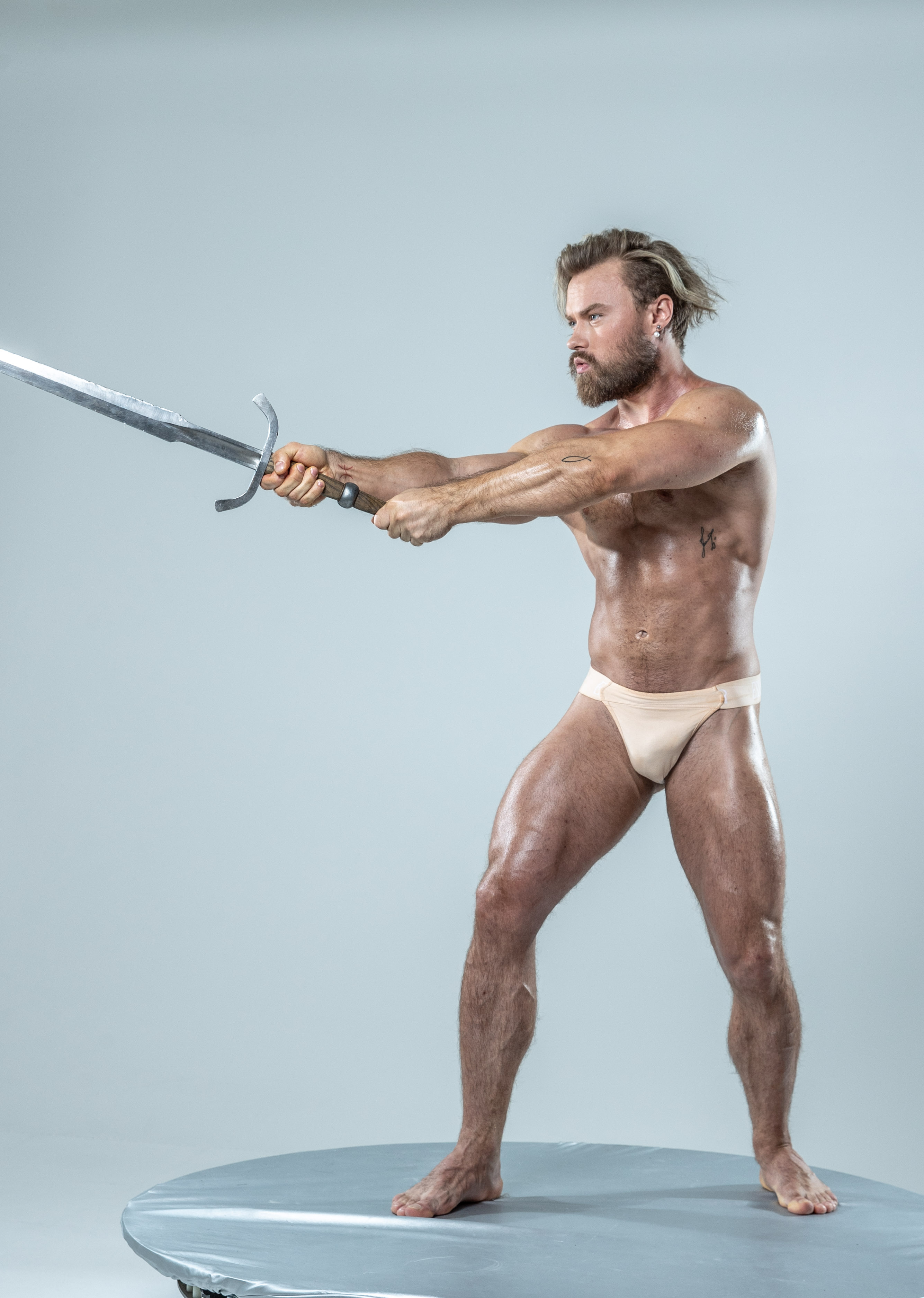 a muscular, barechested man with long blond hair and a beard stands on a circular platform against a light gray background. he holds a large metal sword with both hands, his body tensed as if about to strike. he wears only lightcolored briefs, highlighting his physique. the lighting is soft and diffused, illuminating him evenly. the image evokes a sense of strength and power, reminiscent of a warrior or mythological figure.