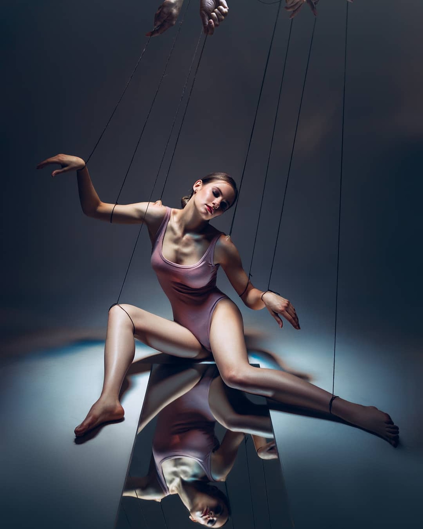 a dramatic image of a young woman posed as a marionette, manipulated by unseen hands and strings. she is sitting on a mirrored surface, her reflection visible below. the overall atmosphere is dark and moody, emphasizing the themes of control and vulnerability.  the woman's pose and expression are both elegant and suggestive of being trapped.  the use of light and shadow creates a powerful visual effect.