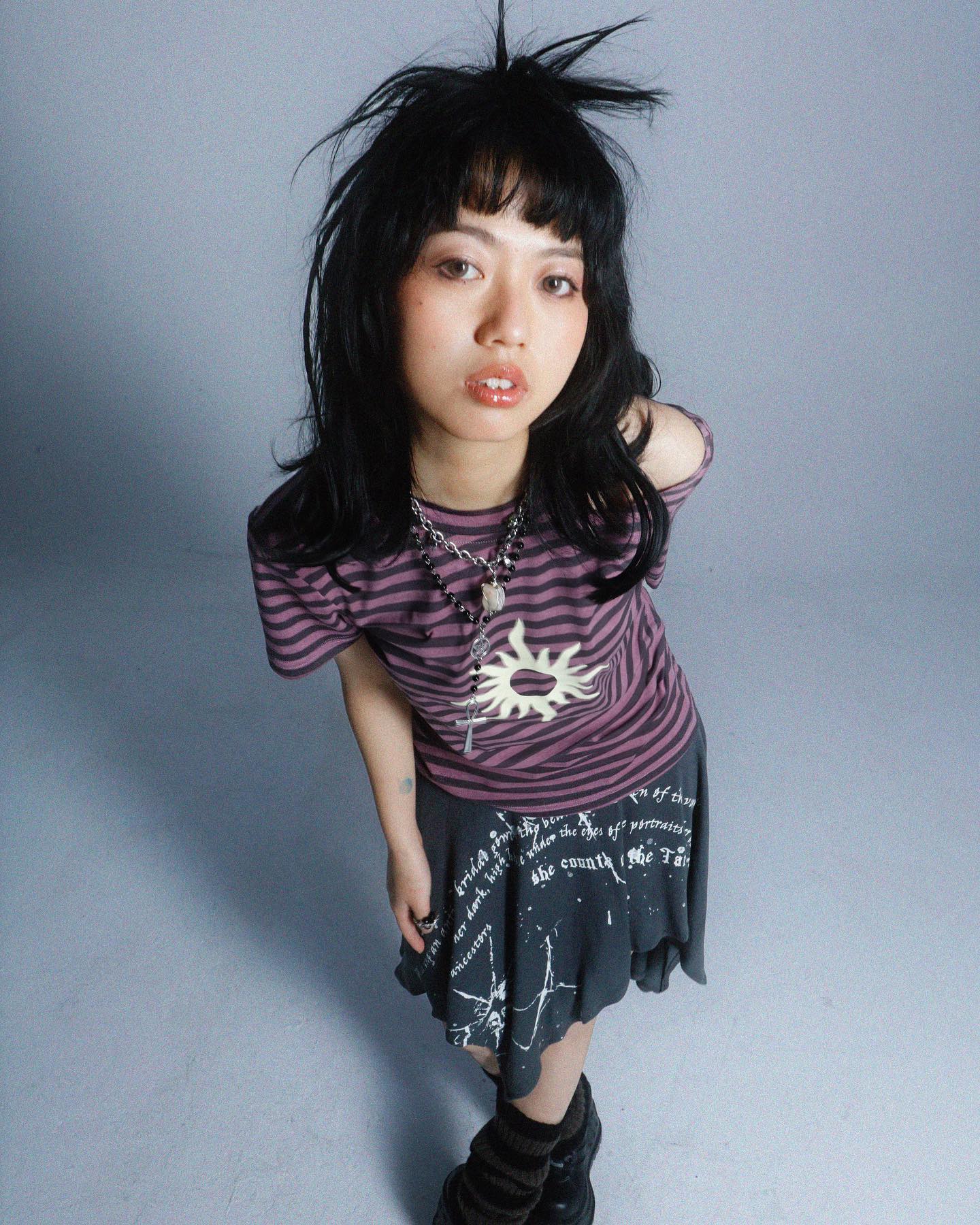 a highangle, full shot of a young asian woman posing against a light grey background. she's wearing a purple striped tshirt with a sun graphic and a black skirt with white text. her dark hair is styled in a messy, slightly spiky look with bangs framing her face. she's wearing dark socks and boots, and has several necklaces on. her overall style is grunge or alternative.  she looks directly at the camera with a slightly serious expression. the image is welllit and has a professional photography quality.