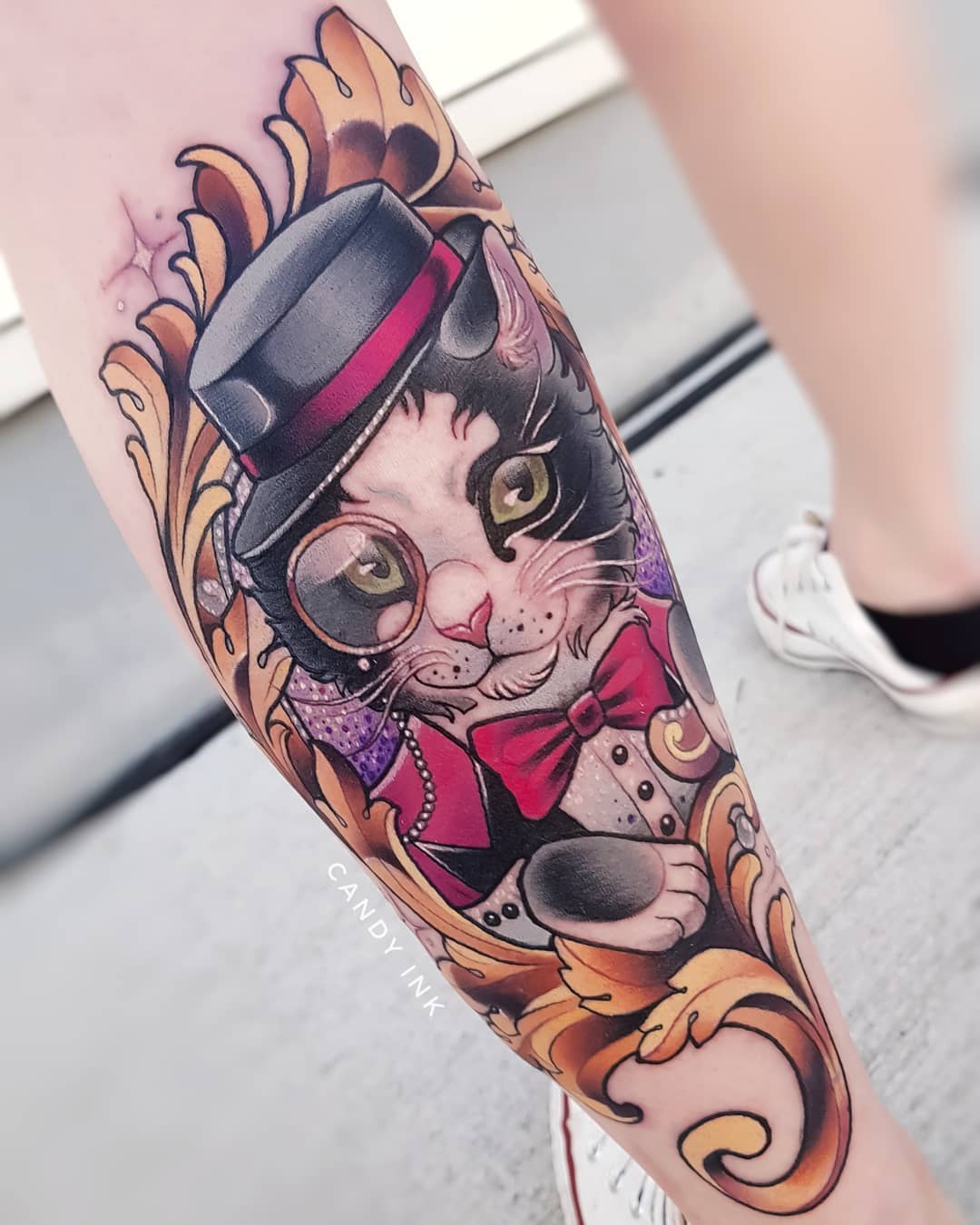 a person's leg with a neotraditional style tattoo of a tuxedo cat wearing a top hat, monocle, and red bowtie. the cat is set within an ornate gold scrollwork design. the tattoo artist's watermark "candy ink" is visible on the image.