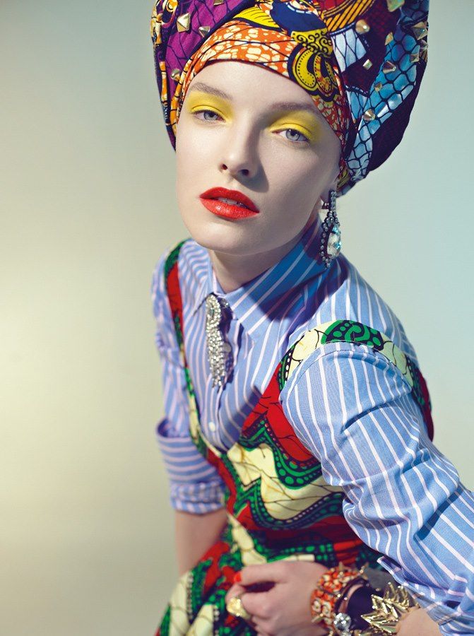 a woman in a colorful african head scarf and patterned dress is looking off camera.  she is wearing blue and white striped shirt, large earrings, and bracelets on her wrist.  her eyes are heavily made up with yellow eyeshadow and black eyeliner.  her lips are a bright red.  the woman is looking downwards as if she is trying to decide what to do next.  the background is a light gray.