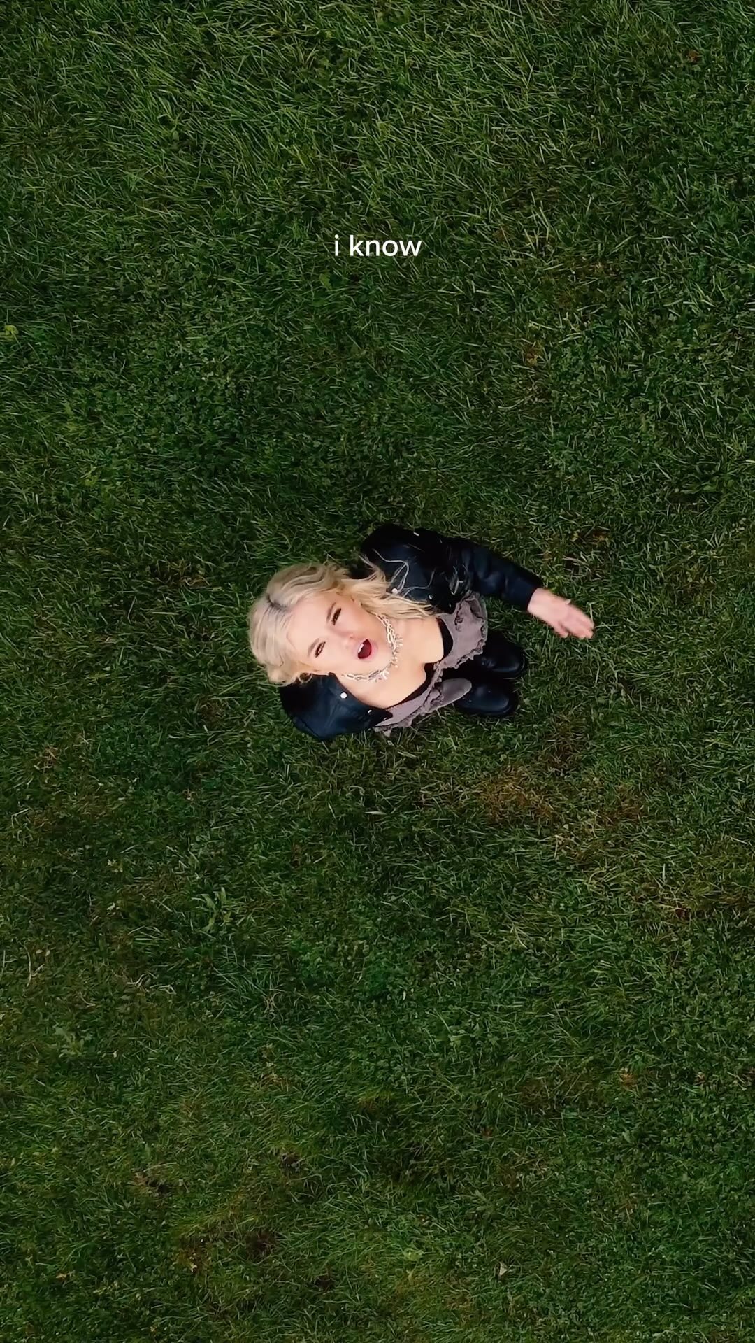 a woman with blonde hair wearing a black jacket and jewelry lies on a patch of green grass and looks up at the camera. the words "i know" are written in white text above her head.
