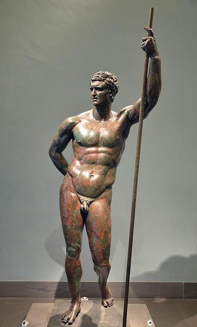 this is a bronze statue of a muscular, nude man standing with his weight shifted onto his right leg.  he holds a spear in his right hand and his left hand rests on his hip.  he has short, curly hair and a serious expression. the statue is displayed in a museum, against a plain gray wall.