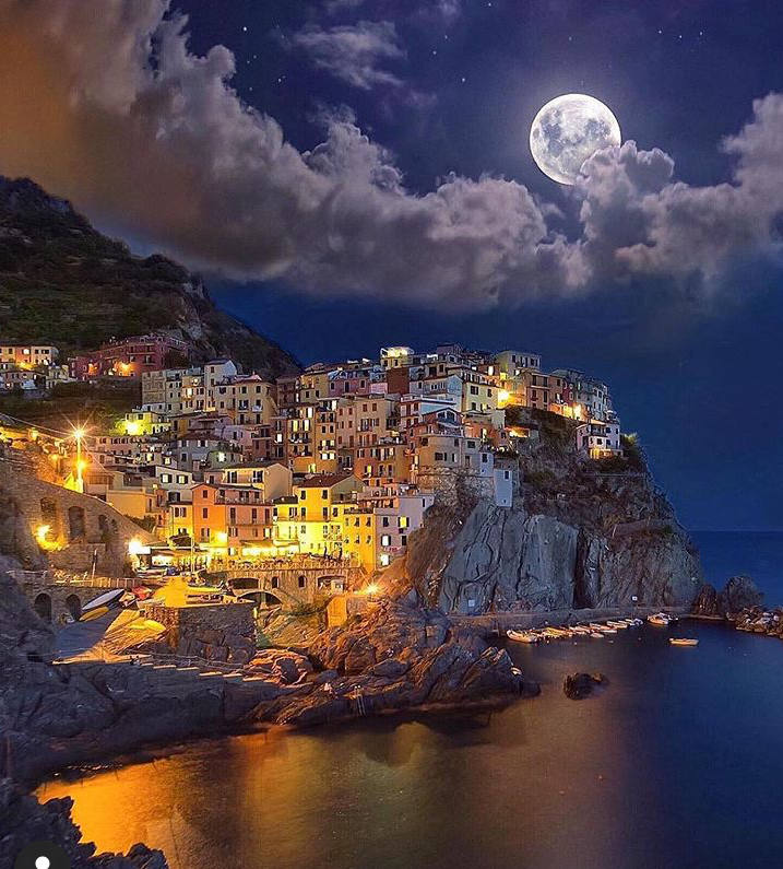 this is a nighttime landscape image of manarola, cinque terre, italy. the village, with its colorful buildings, is perched on a cliff overlooking the sea. the coastline is rocky, and there is a small harbor with boats. lights illuminate the buildings and surrounding areas. a full moon is prominent in the sky, surrounded by clouds.