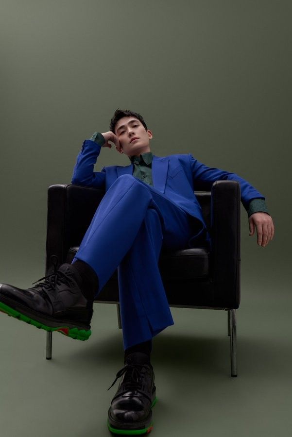 a man in a blue suit with a green shirt is sitting in a black chair. his left foot is on the ground, his right foot is bent and raised with his shoe showing. he is looking at the camera. the background is a green wall.
