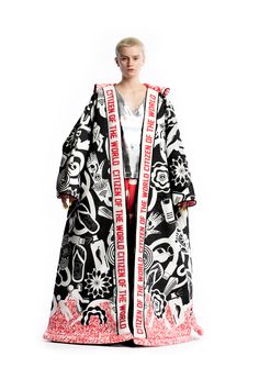 a woman wearing a long black, white, and red patterned coat with "citizen of the world" text repeated down the front is looking at the camera. the background is a white studio space.
