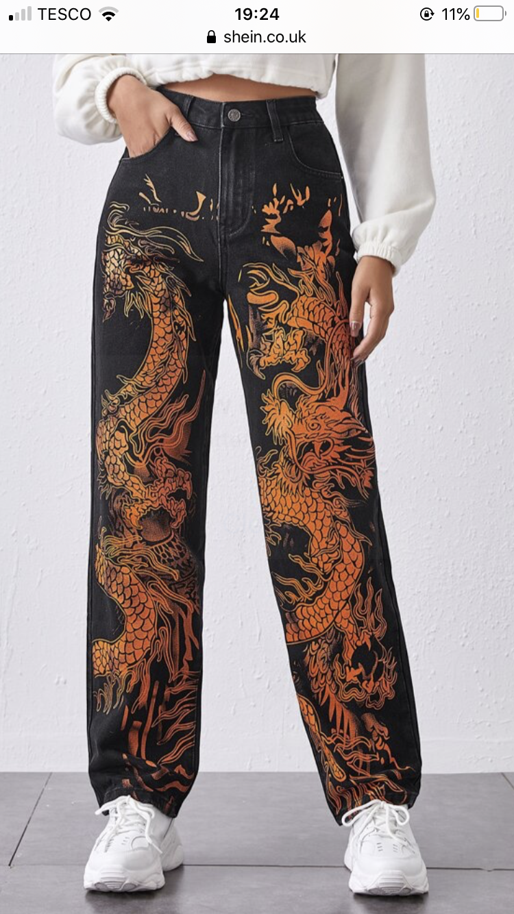 a woman is modeling a pair of black jeans with a large orange and yellow dragon print. she is standing in front of a plain background, and the jeans are the main focus of the image. the jeans are highwaisted and straightlegged, and the woman is wearing them with a white top.