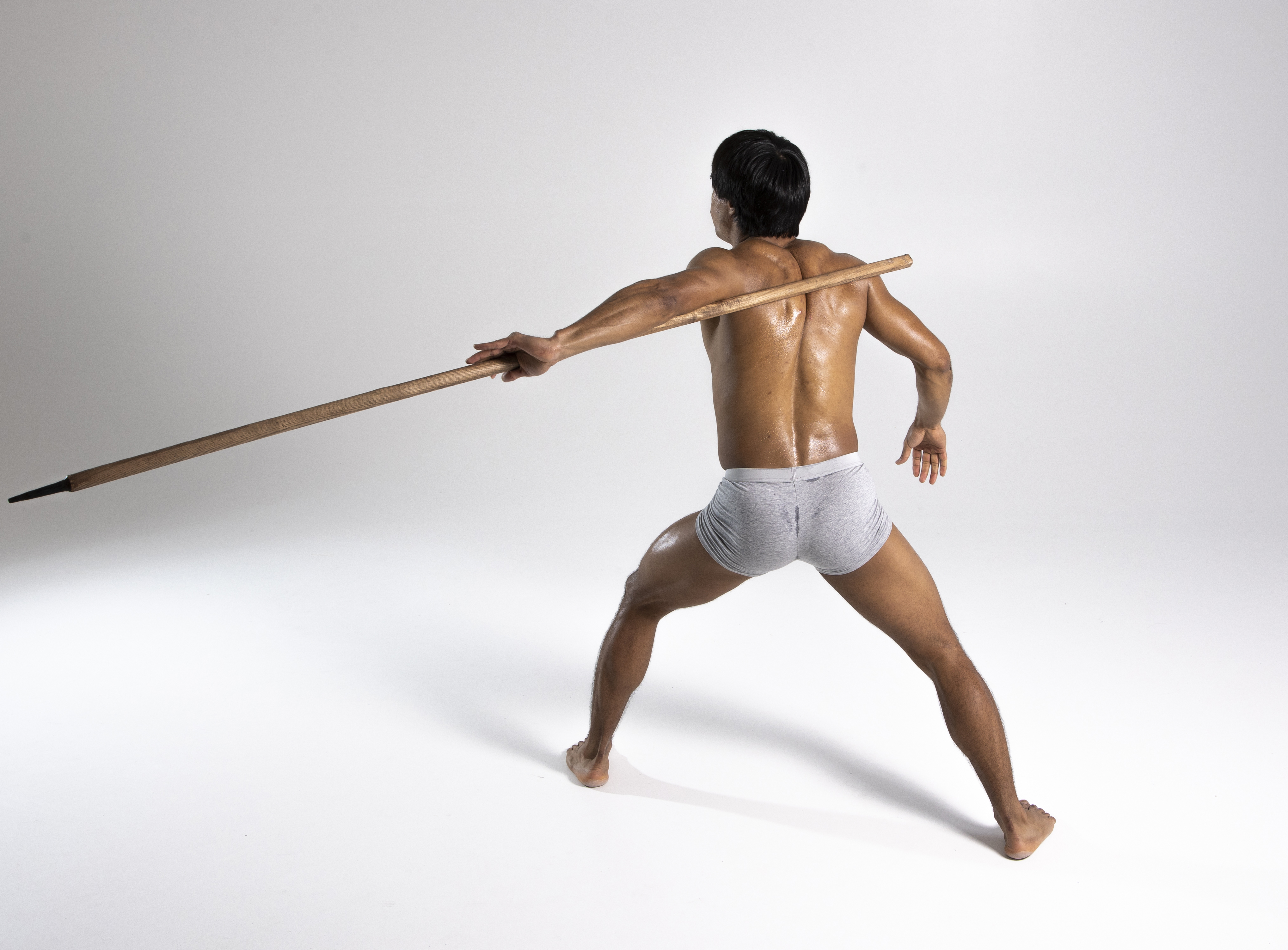 a muscular, shirtless man is standing barefoot in a studio, holding a spear across his back with one hand extended. he is wearing grey shorts and his back is facing the viewer. the background is plain white. the man appears to be in a ready stance, possibly for martial arts.