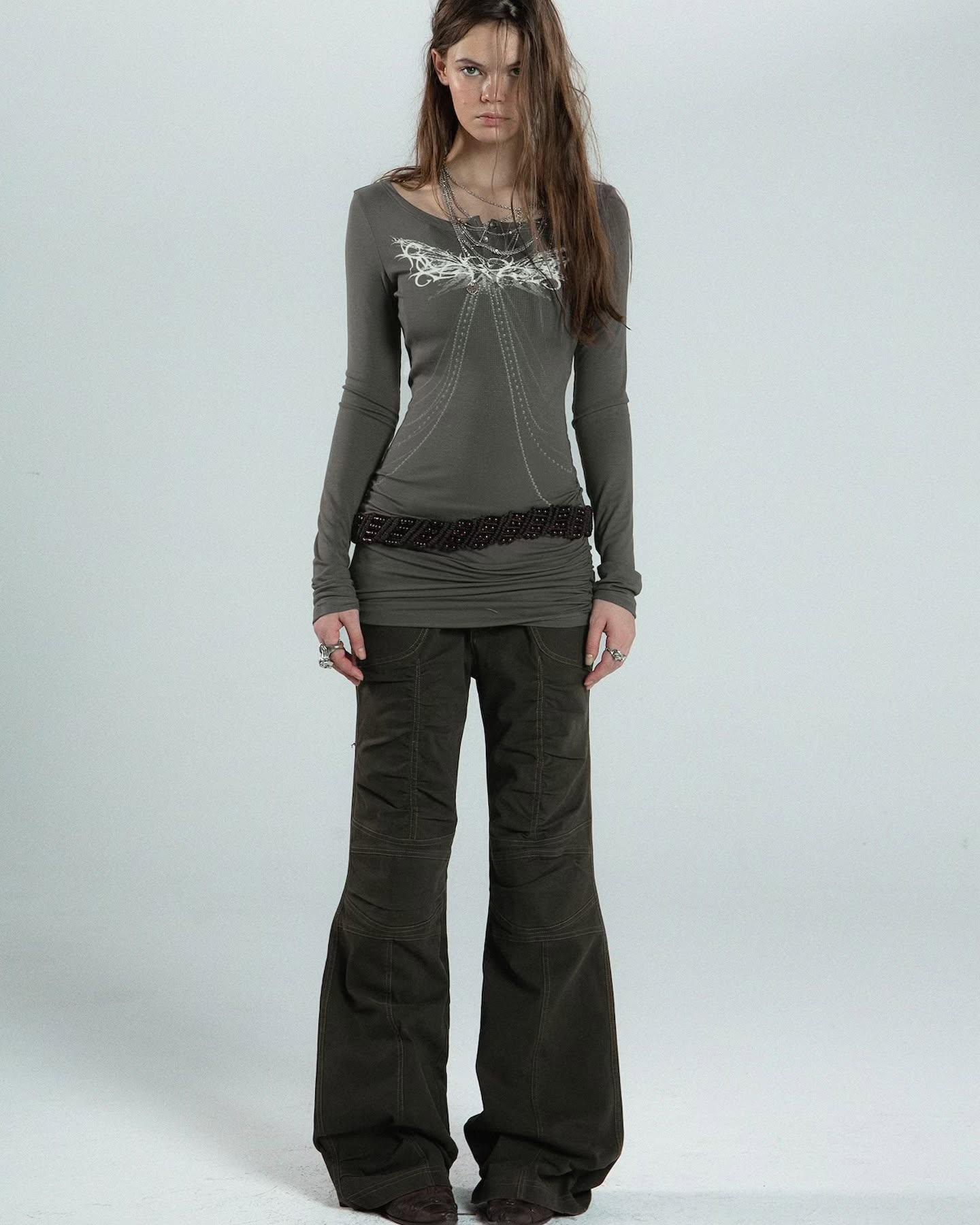 a fullbody shot of a young woman posing against a light grey background. she is wearing a longsleeved, grey shirt with a subtle white design on the chest, dark green bellbottom pants, and a dark brown beaded belt. the model has long, straight brown hair and is wearing minimal jewelry including a necklace.  her pose is relaxed and confident, showcasing the clothing. the image is welllit and professionally taken, possibly for a fashion catalog or website.