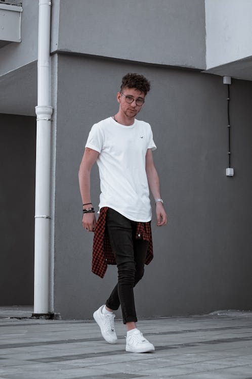a young man with curly hair and glasses is walking outdoors in front of a gray building. he is wearing a white tshirt, black jeans, a red plaid shirt tied around his waist, and white sneakers. his style is casual and trendy. the image is a full body portrait, and the setting is urban.