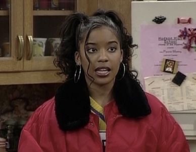 a young africanamerican woman with long, curly black hair wearing a red jacket with a black fur collar, is smiling and looking at the camera.