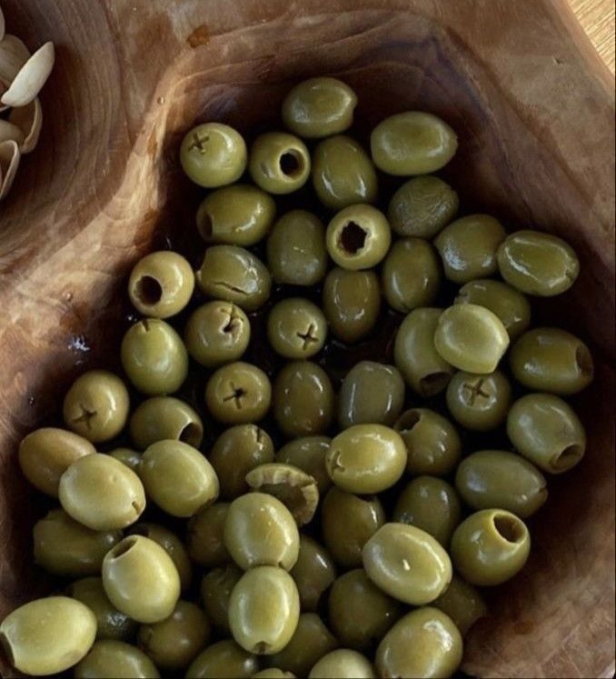 a closeup shot of a bowl filled with green pitted olives. the olives are various shades of green and some have a cross cut in the top, some have a hole in the middle. the bowl appears to be made of wood. there are some pistachio shells to the left.