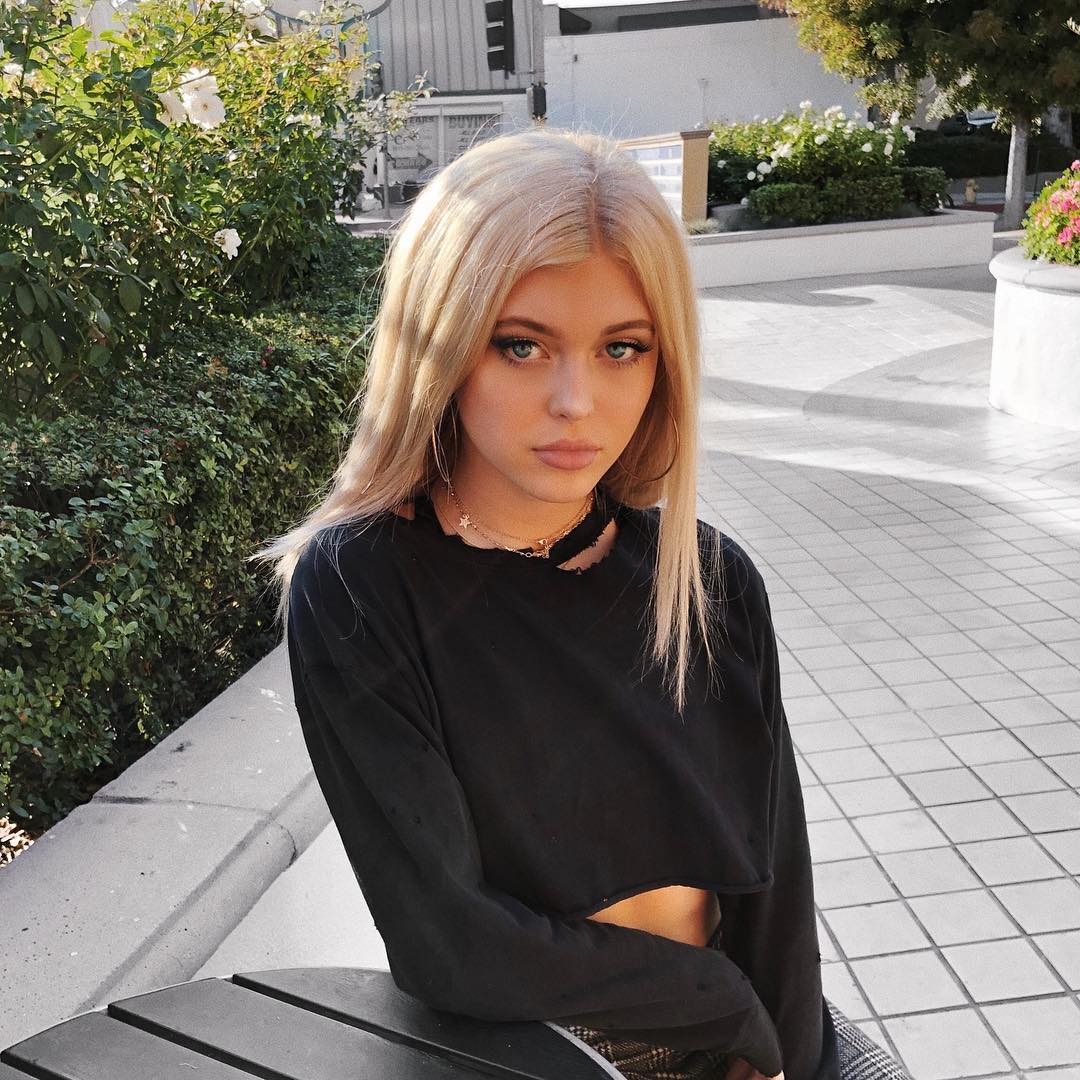 a young woman with blonde hair and blue eyes poses outdoors. she is wearing a black longsleeved crop top with a distressed neckline. she accessorizes with a necklace and hoop earrings. the background includes pavement, greenery, and shadows, suggesting an urban setting.