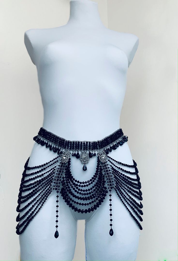 a mannequin is wearing a decorative black beaded waist belt. the belt is wide and adorned with intricate beadwork, featuring dangles of varying lengths that cascade down the mannequin's hips. the beads are black, creating a statement piece. the mannequin is a pale color, providing a contrast to the dark beads.