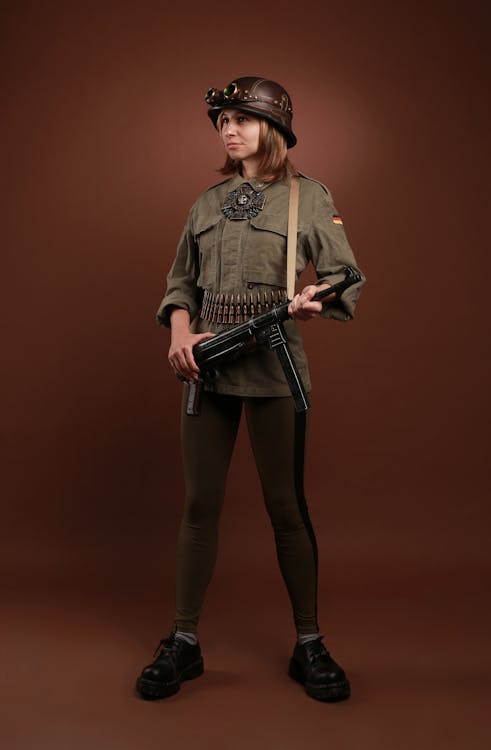 a fullbody studio shot of a woman dressed in a steampunkinspired military uniform. she's wearing a brown leather helmet with goggles, a green military jacket with a german flag patch and ammunition belt, and black leggings with black boots. she's holding an mp40 submachine gun. the background is a simple brown backdrop.  the overall style blends historical military elements with steampunk aesthetics.
