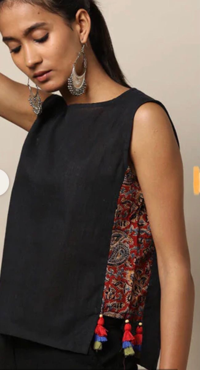 a woman poses while wearing a sleeveless black top with a slit on the side that reveals a patterned fabric underneath, and tassels hanging off the bottom. she has silver earrings.