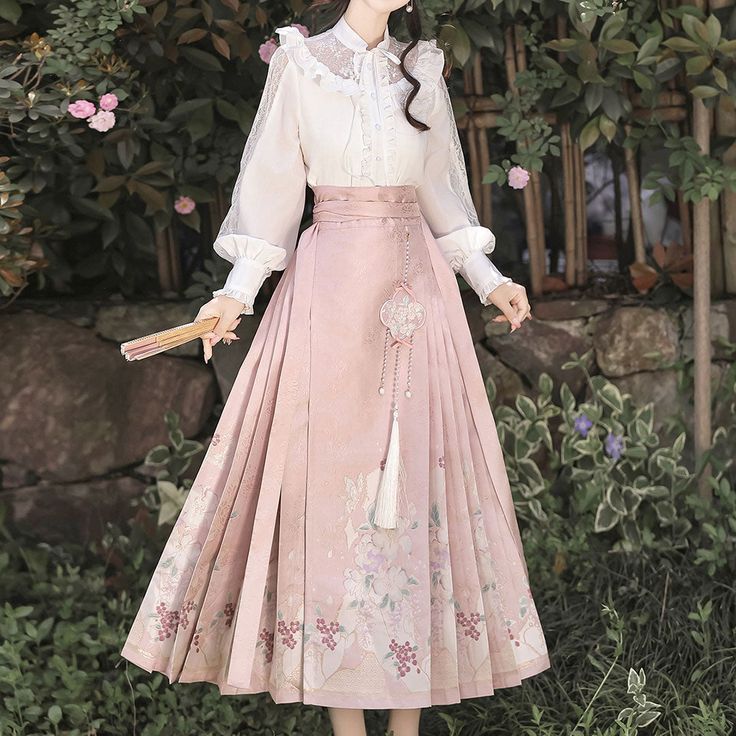 a woman stands in a garden wearing a pink floral print skirt with a white lace blouse. the skirt is long and flowing, with a high waist and a pleated design. the blouse has long sleeves and a ruffled collar. the woman is holding a fan in her right hand. the garden is lush and green, with flowers in the background. the lighting is soft and diffused, creating a dreamy and romantic atmosphere.