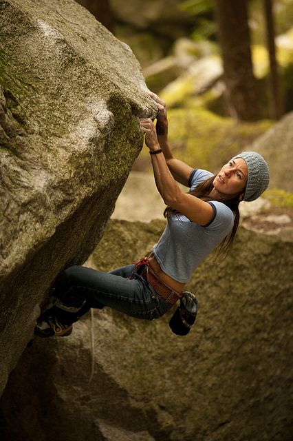 a woman is bouldering in a forest. she is wearing a blue tank top, jeans, and a grey beanie. she has her hand on a large boulder and is looking up with a determined expression. the lighting is natural and dappled, creating a sense of depth and texture. the image conveys a sense of strength, athleticism, and the beauty of the natural world.