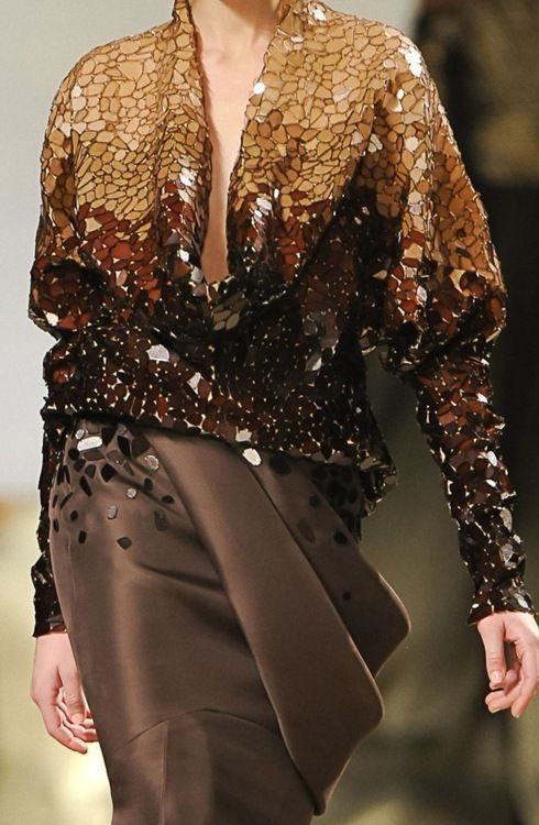 a woman model walks the runway in a brown sequined dress with a dramatic drape and long sleeves. the dress is elegant and stylish. the model is posed in a graceful manner, highlighting the dress's unique design.