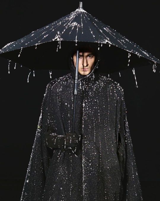 a man stands against a black background holding a black umbrella adorned with water droplets or beads. the umbrella is open and centered over his head. he is wearing a black, hooded cape that is also embellished with beads or droplets, giving the impression of rain. he has a serious expression and is wearing black gloves. the overall aesthetic is dark and dramatic.