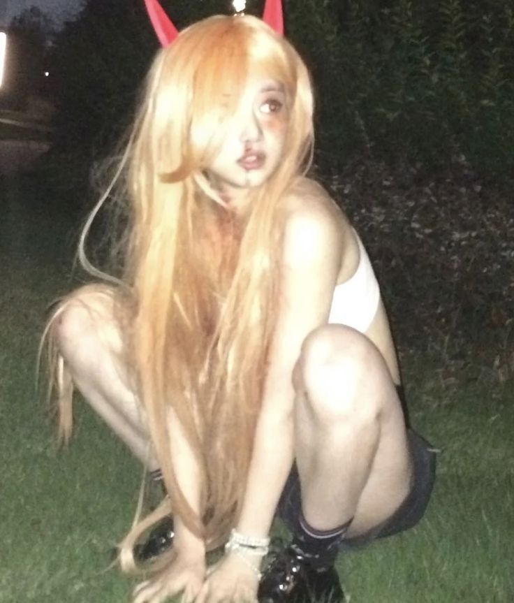 a young woman with long blonde hair and red horns sitting on grass wearing a white top and black shorts. she looks like she's posing for a picture in the dark, outdoors.