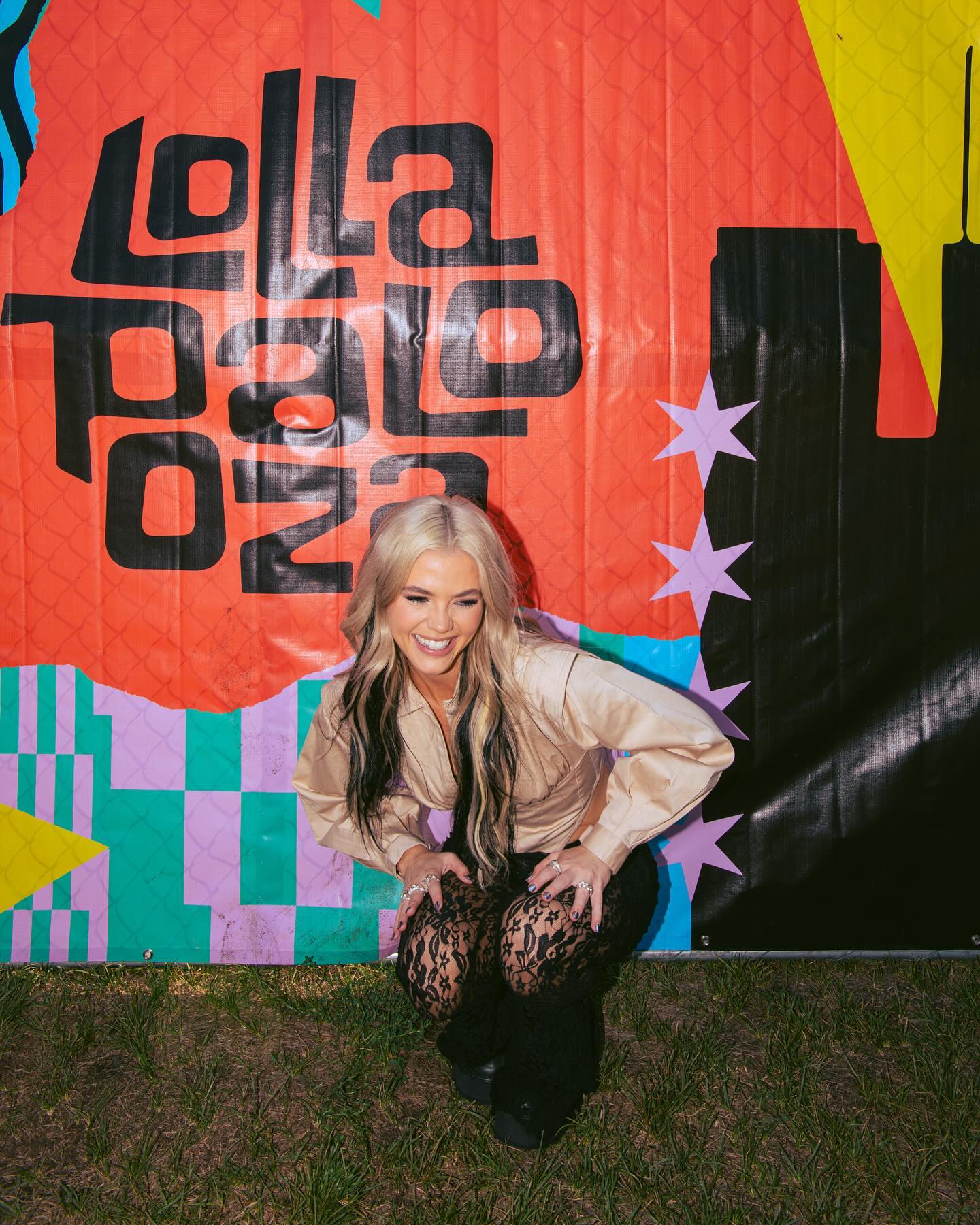 a smiling woman with blonde hair and black highlights is crouching in front of a lollapalooza backdrop. she is wearing a beige top and black lace pants. the background includes a colorful banner and green grass.