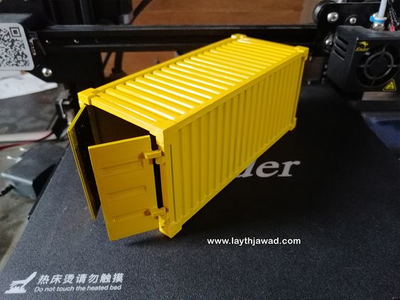 a yellow shipping container model that has been 3d printed sits on a black surface with a white text sticker on it.  a 3d printer is in the background.