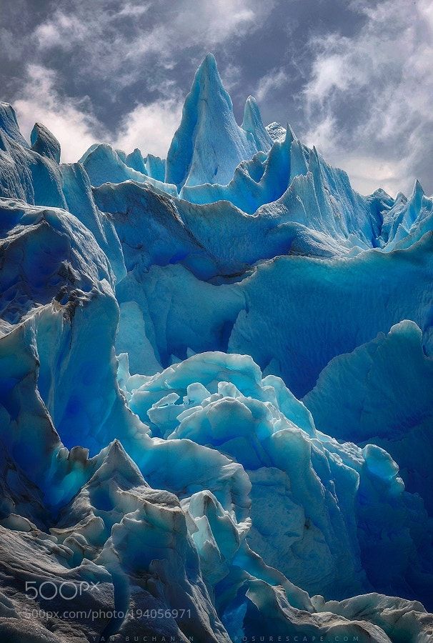 the image showcases a stunning, closeup view of a glacier. the dominant colors are various shades of blue and white, creating a cold and icy atmosphere. the glacier's surface is jagged and uneven, with sharp peaks and deep crevices, revealing its age and formation. overlapping ice layers and formations create interesting textures and patterns. the sky above is cloudy, adding drama to the scene. the photograph focuses primarily on the intricate details of the glacier itself.