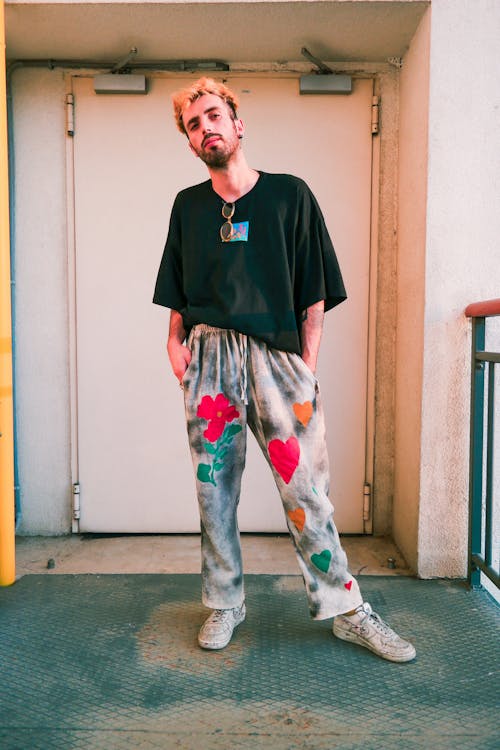 a young man with blonde hair is standing in a doorway, posing for a photo. he's wearing a black tshirt, grey tiedye pants with painted flowers and hearts, and white sneakers. his style is casual and streetwearinspired. the background is a simple urban setting with a concrete floor and a door. the overall mood is relaxed and fashionable.