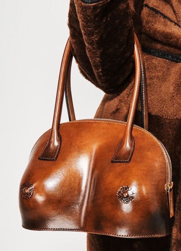 a brown leather handbag shaped like breasts is held by its handles. the bag has a rounded shape with two raised areas resembling breasts and textured embellishments resembling nipples. a zipper closure runs across the top. the bag is held against a person wearing a brown, furry coat.