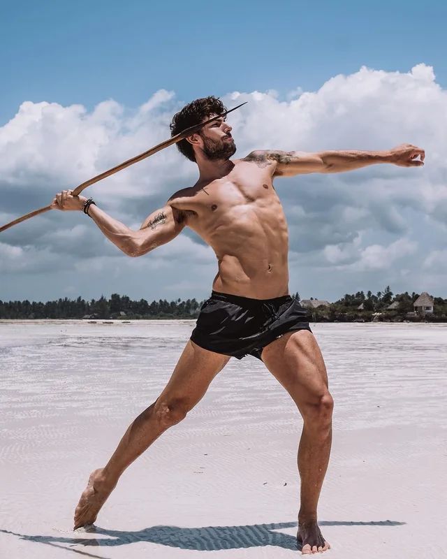 a handsome, muscular, tanned man stands on a white sandy beach, throwing a javelin with a powerful stance. he's wearing black swim shorts. the sky is blue with white clouds and the water is calm and clear. he looks determined and ready for action. the image evokes a sense of strength, athleticism, and summer vacation.