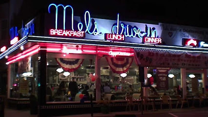 a photo of mel's drivein diner at night. the diner is lit up with neon signs advertising "breakfast lunch dinner". there are people sitting inside at tables and a few people standing. the exterior of the building is decorated with red, white, and blue bunting.