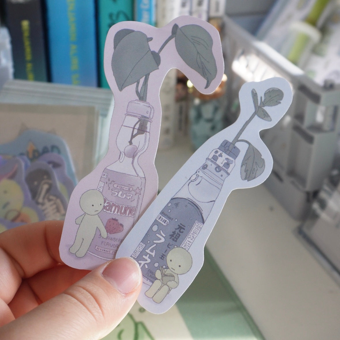 two stickers of ramune soda bottles with plants growing out of them. the stickers are both very cute and illustrated in a cartoon style. one sticker is of a ramune soda bottle with a green plant and the other is of a dark colored ramune soda bottle with a purple plant. both stickers have a small cartoon character next to the bottle. the stickers are on a white background.