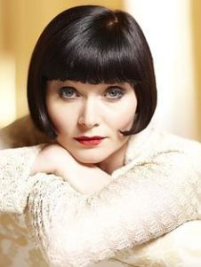 a closeup portrait of a woman with short black bobbed hair and bangs. she is wearing a white sweater and has a serious expression on her face. her lips are painted red. she is looking directly at the camera.