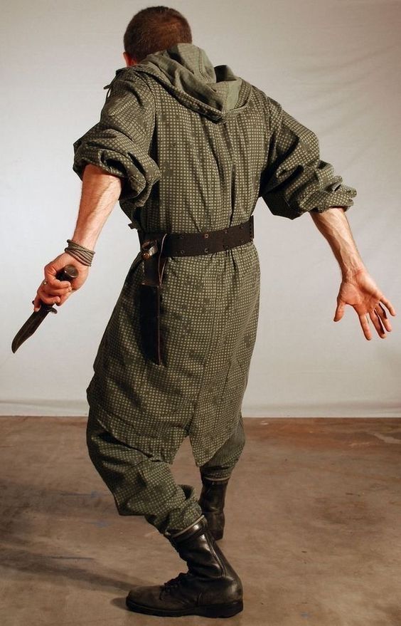 a man in a camouflage military jacket and boots is standing with his back to the camera holding a knife in his right hand. he's looking down at his weapon with a serious expression. he is wearing a leather belt with a sheath on the right side. the background is a simple white wall.