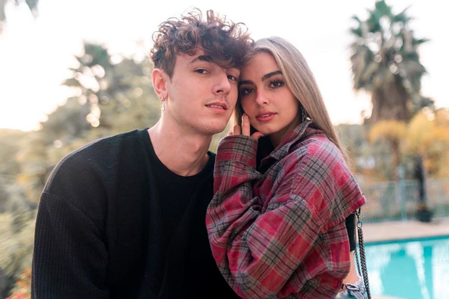a young couple stands close together outdoors, the man is wearing a black sweater and the woman a red plaid shirt. they look into each other's eyes and are smiling. they appear to be very much in love. the background is blurry and out of focus, with a swimming pool and palm trees visible. the image evokes a sense of romance and happiness.